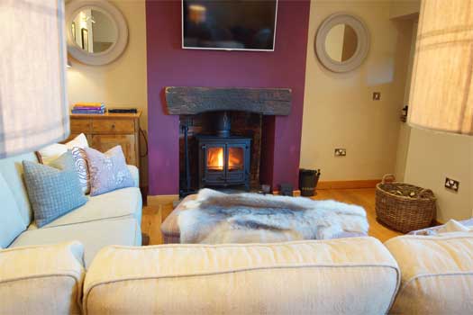 Cosy wood-burning stove at The Cottage set into an aubergine feature wall with fur throw and cushions