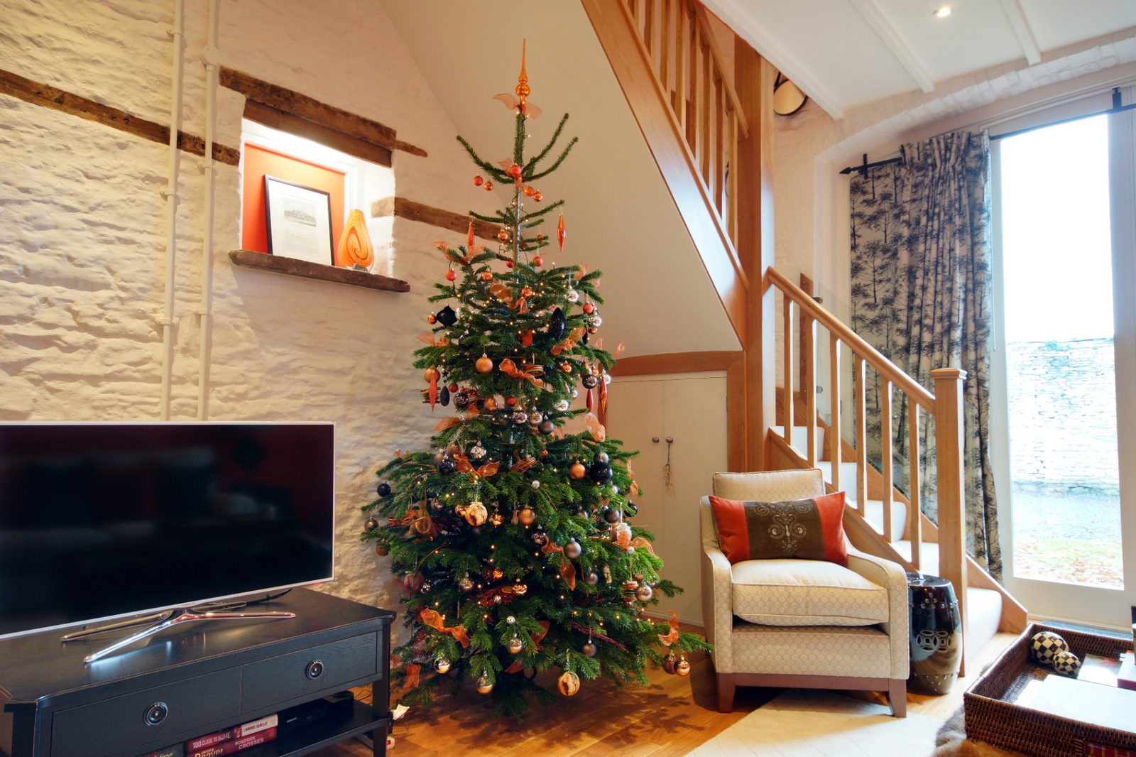 A Christmas tree trimmed with copper and bronze baubles beside a vaulted stone-walled sitting room with oak staircase at Ty Mawr