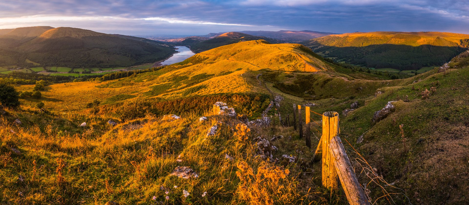 Golden hour light over the Brecon Beacons with a reservoir nestled between glowing hillsides and a wooden fence in the foreground