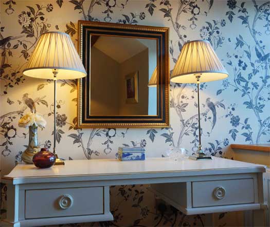 Detail of the bedroom dresser at The Cottage with paired lamps and gilt-framed mirror on floral wallpaper