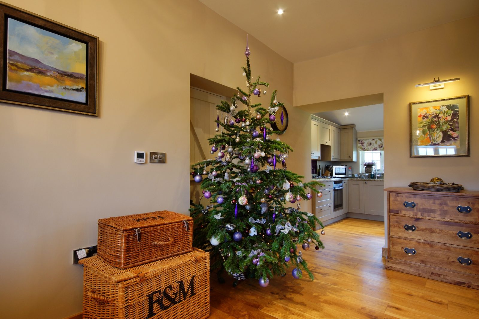 A tall Christmas tree decorated in purple and silver baubles beside wicker Fortnum and Mason hampers in a Ty Mawr cottage hallway
