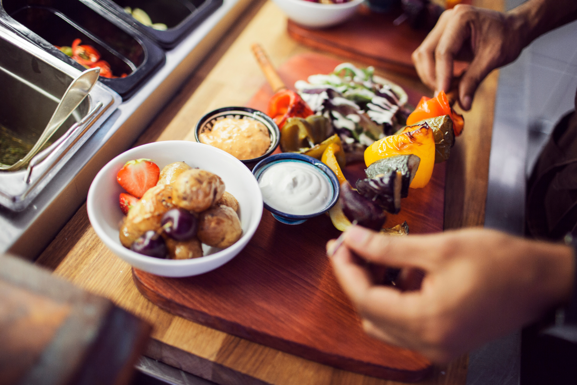 Chef plating colourful skewers of grilled peppers and courgette with new potatoes, strawberries and dips on a wooden board