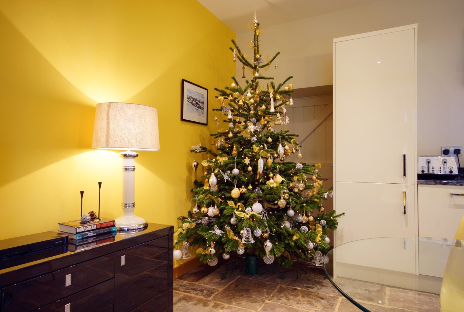 A gold and silver decorated Christmas tree against a warm yellow wall in a beamed Ty Mawr cottage kitchen